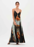 A model wears a floor-length spaghetti-strap maxi dress with a black bodice and a flowing skirt featuring a multicolor animal and landscape print on a light blue-green background, set against a plain white studio backdrop.